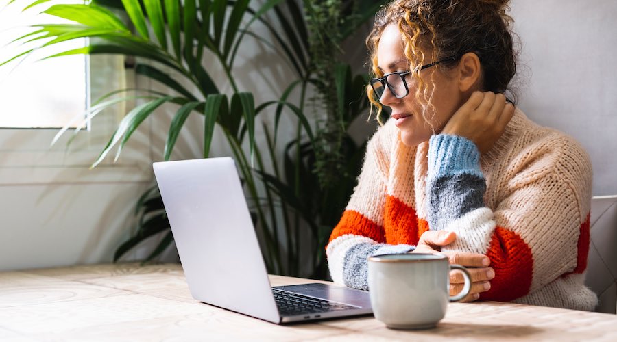 A woman sitting at a table at home, reading a laptop screen.